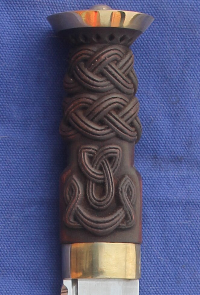 Dirk has brass cap and collar above blade. Grip is carved with 2 three band pleat knotwork bands and 2 knots on the shoulder above the blade.